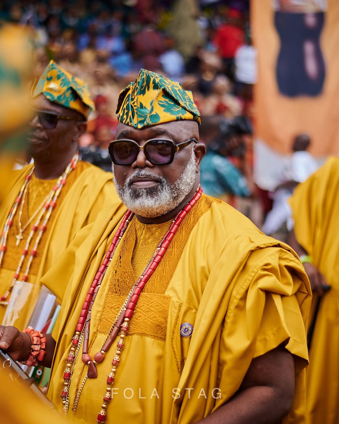 Close-up portrait of an attendee with a grey beard at Ojude Oba wearing a mustard yellow embroidered Agbada, a matching patterned Fìlà cap, sunglasses, and coral beads.