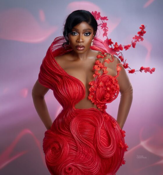 Actress Bolaji Ogunmola posing in a structured red avant-garde gown with sculptural piping, a sheer bodice, and a 3D red floral branch extending over her shoulder.
