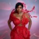 Actress Bolaji Ogunmola posing in a structured red avant-garde gown with sculptural piping, a sheer bodice, and a 3D red floral branch extending over her shoulder.