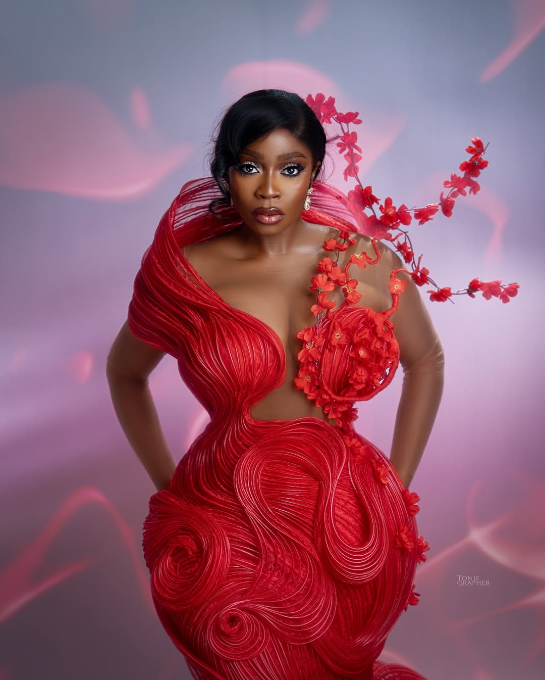Actress Bolaji Ogunmola posing in a structured red avant-garde gown with sculptural piping, a sheer bodice, and a 3D red floral branch extending over her shoulder.