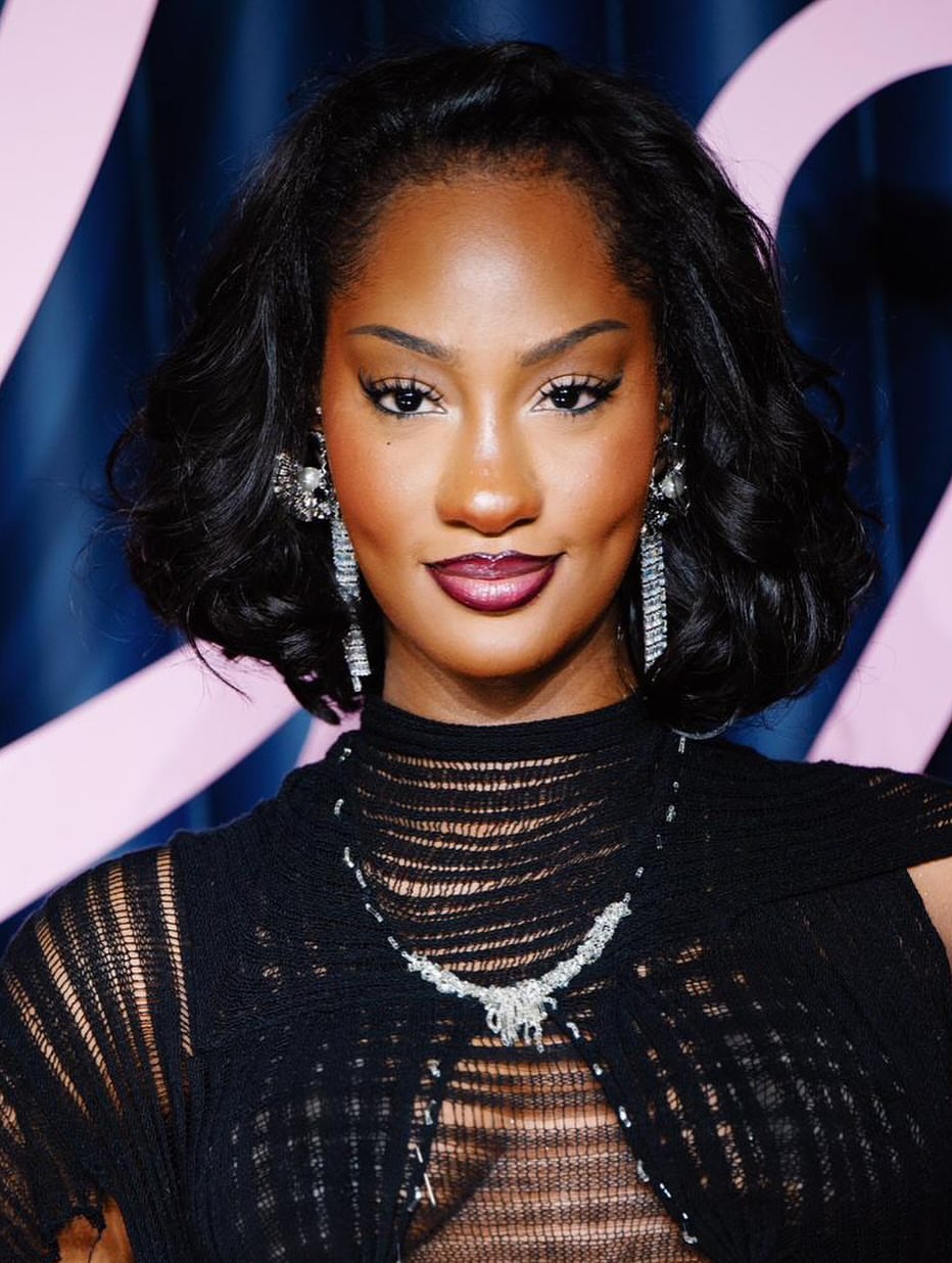 close-up red carpet portrait of Nigerian singer Tems featuring a voluminous wavy bob hairstyle, a dramatic winged eyeliner look, and a deep glossy ombré lip, wearing a textured black high-neck gown with silver jewelry.