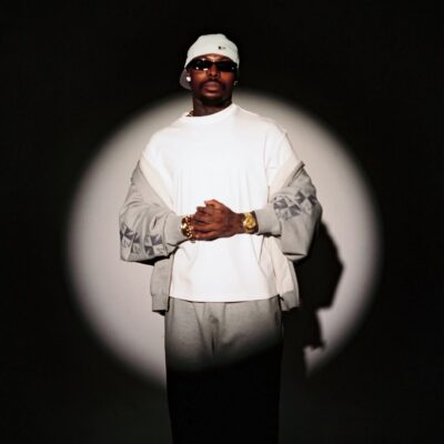 Asake posing in a spotlight wearing a white t-shirt and grey tracksuit for his MONEY album campaign.