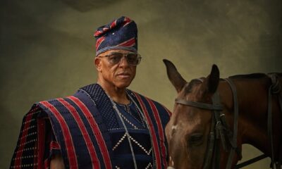 Wide shot of Farooq Oreagba in a striped prestige Agbada holding a gold-handled cane while standing beside a saddled brown horse against an olive background.