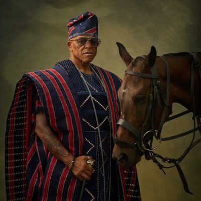 Wide shot of Farooq Oreagba in a striped prestige Agbada holding a gold-handled cane while standing beside a saddled brown horse against an olive background.