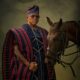 Wide shot of Farooq Oreagba in a striped prestige Agbada holding a gold-handled cane while standing beside a saddled brown horse against an olive background.