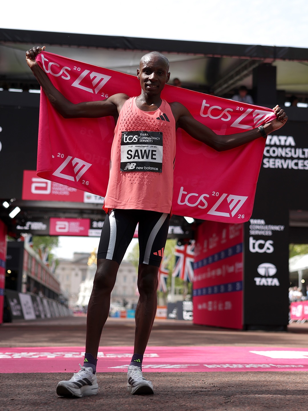 abastian Sawe standing at the finish line of the 2026 London Marathon holding a pink TCS event flag after breaking the sub-two-hour world record.
