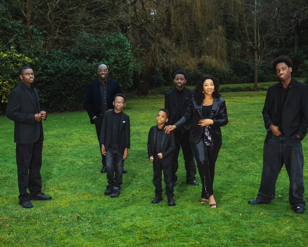 The Iyiegbu family, including Obi Cubana and Ebele Iyiegbu, standing on a lush garden lawn in coordinated all-black formal attire.
