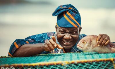 Lead actor William Lamptey as retired fisherman Atta Oko, wearing a traditional blue and orange patterned cap, smiling and pointing next to a large fish on a green fishing net.