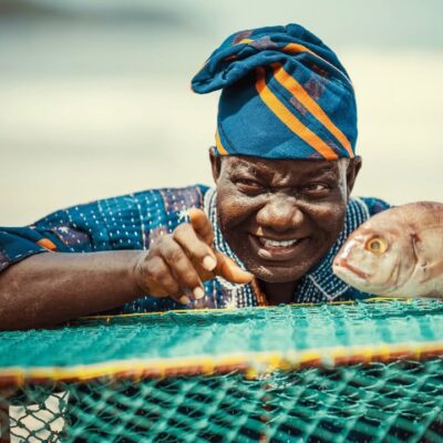 Lead actor William Lamptey as retired fisherman Atta Oko, wearing a traditional blue and orange patterned cap, smiling and pointing next to a large fish on a green fishing net.