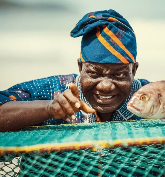 Lead actor William Lamptey as retired fisherman Atta Oko, wearing a traditional blue and orange patterned cap, smiling and pointing next to a large fish on a green fishing net.
