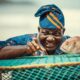 Lead actor William Lamptey as retired fisherman Atta Oko, wearing a traditional blue and orange patterned cap, smiling and pointing next to a large fish on a green fishing net.