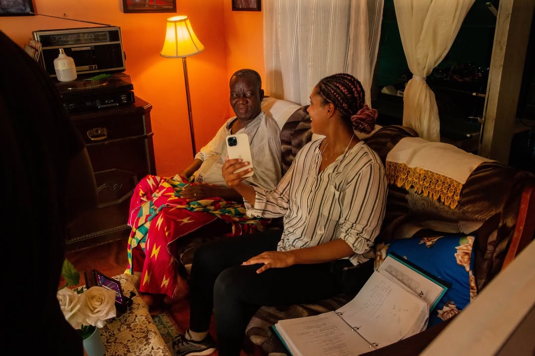 Ghanaian-American director Zoey Martinson holding a phone while sitting on a couch next to lead actor William Lamptey during a behind-the-scenes script review session for the film The Fisherman.
