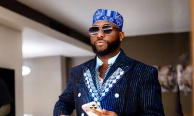 Close-up of Afrobeats star Davido in a navy pinstripe suit with a checkered lapel, blue traditional cap, and sunglasses while holding a phone.