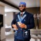 Close-up of Afrobeats star Davido in a navy pinstripe suit with a checkered lapel, blue traditional cap, and sunglasses while holding a phone.