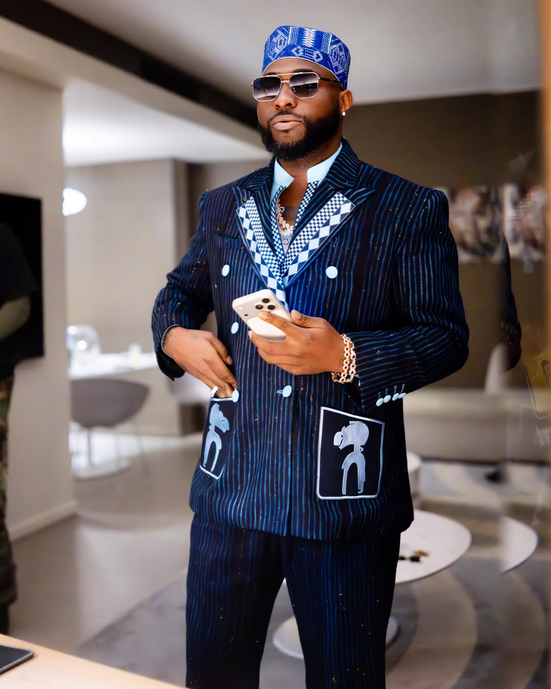 Close-up of Afrobeats star Davido in a navy pinstripe suit with a checkered lapel, blue traditional cap, and sunglasses while holding a phone.