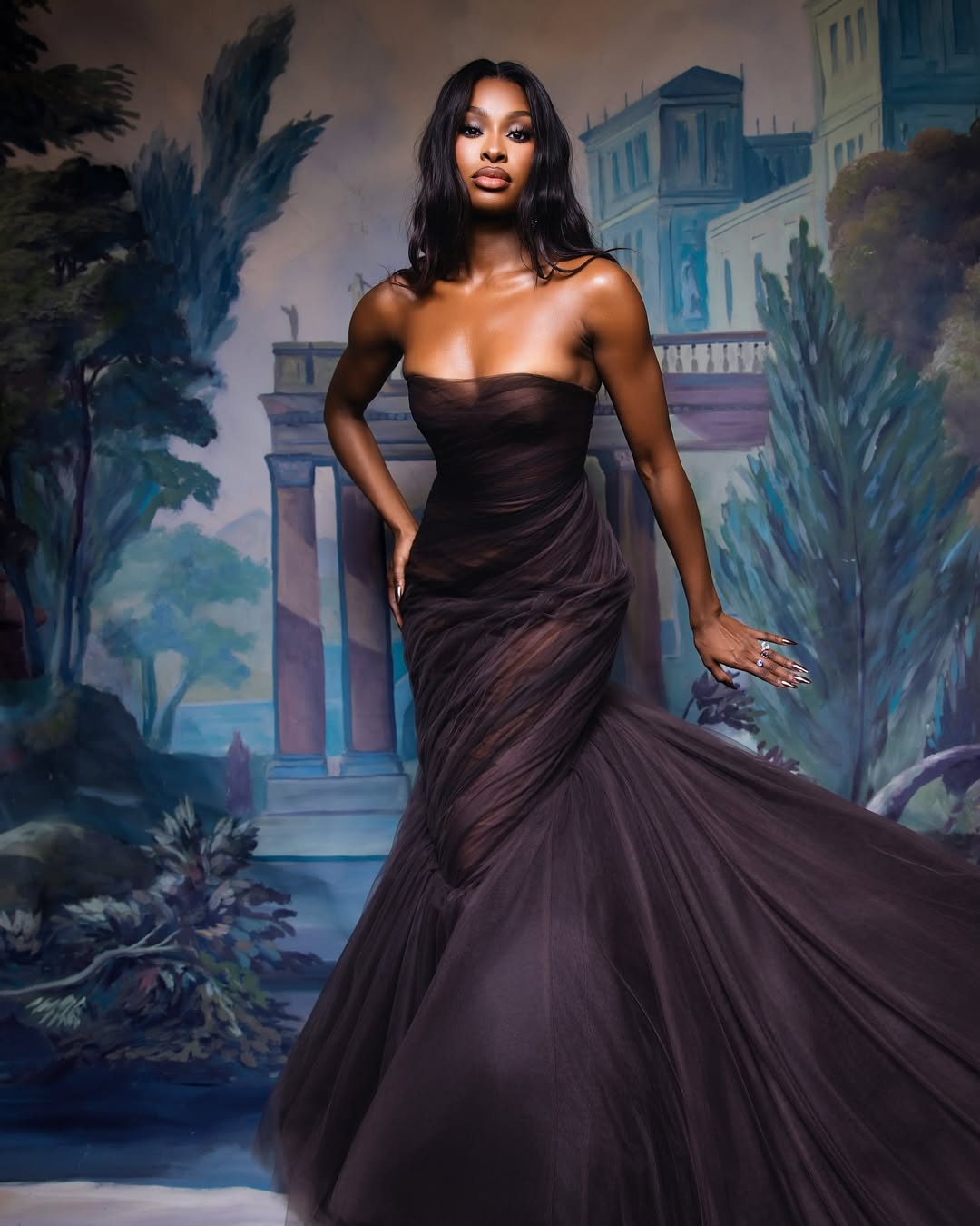 Actress and singer Coco Jones attends the 2026 Met Gala in a custom aubergine (deep purple) sheer tulle gown by Prabal Gurung. The look features a classic mermaid silhouette and monochromatic soft waves styled to match the gown's specific purple hue.