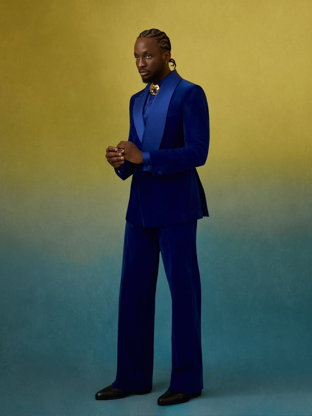 Actor Chimezie Imo posing against a gradient yellow-to-teal backdrop in a double-breasted electric blue velvet suit by Fai World with a gold statement brooch.