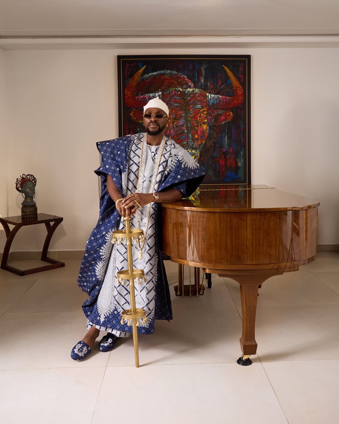 Tobi Bakre leaning on a grand piano in a custom blue and white monogram canvas denim Agbada by Deji and Kola, holding a traditional Șaworoide staff.