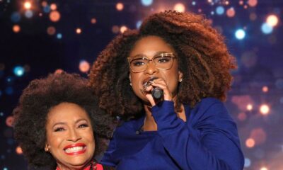Hollywood legend Jenifer Lewis in a sheer black jacket over a red turtleneck smiling with her arm around talk show host Jennifer Hudson, who is holding a microphone and wearing a blue top with tan trousers on stage.