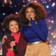 Hollywood legend Jenifer Lewis in a sheer black jacket over a red turtleneck smiling with her arm around talk show host Jennifer Hudson, who is holding a microphone and wearing a blue top with tan trousers on stage.
