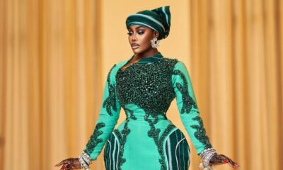 Mid-shot of Nana Akua Addo wearing a green avant-garde gown with architectural hip padding, holding dramatic silver chandelier kalire hanging from stacked wrist cuffs.