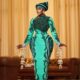 Mid-shot of Nana Akua Addo wearing a green avant-garde gown with architectural hip padding, holding dramatic silver chandelier kalire hanging from stacked wrist cuffs.