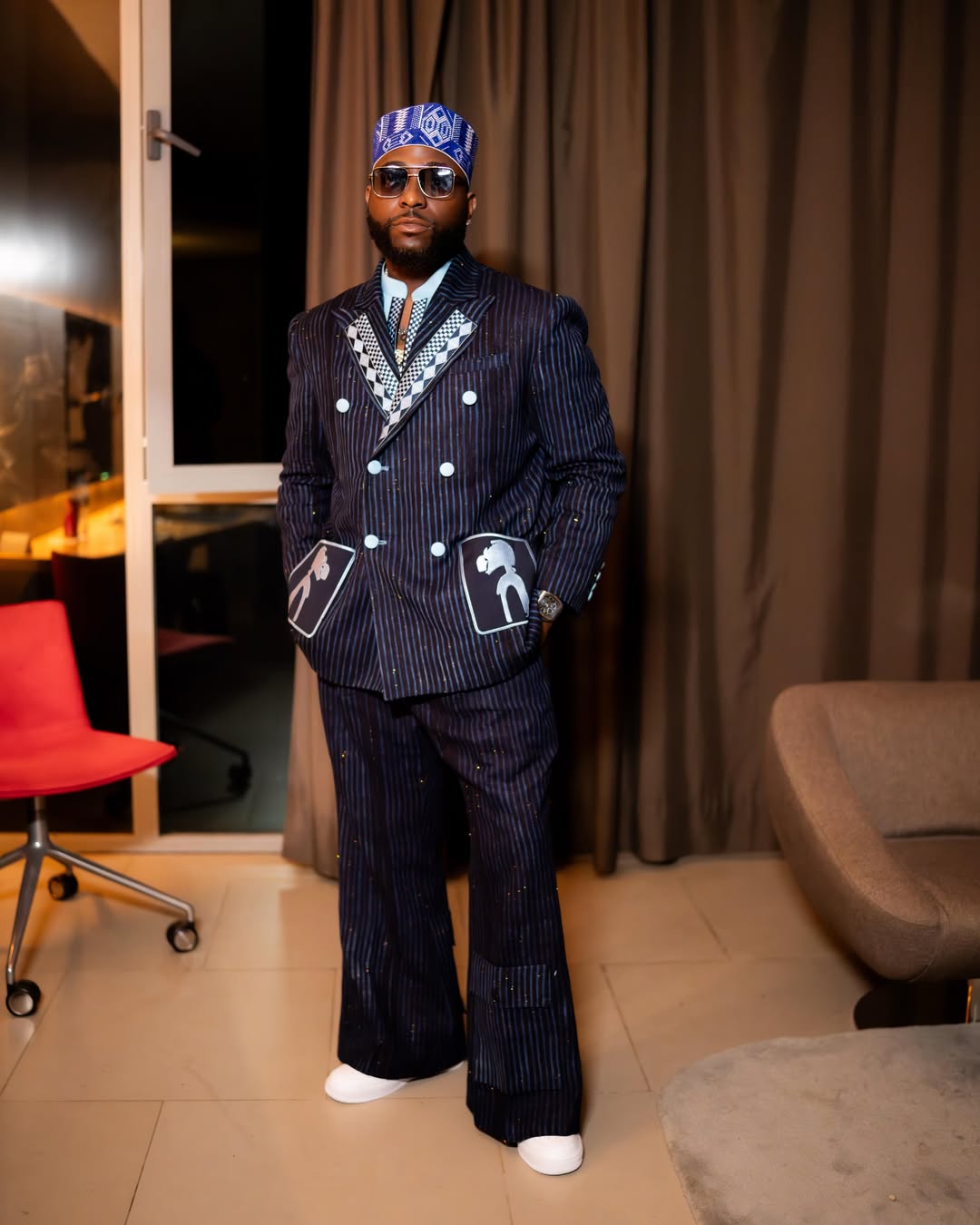 Full-length portrait of Davido standing indoors in a custom navy blue pinstripe double-breasted suit, matching traditional cap, and white sneakers.