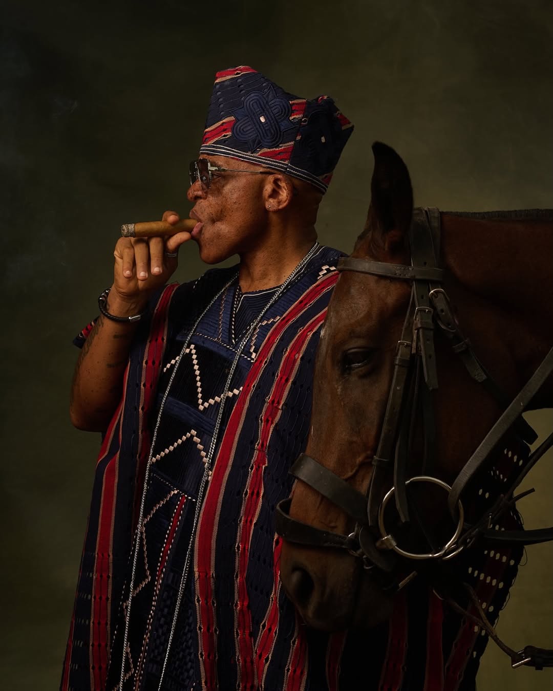 Close-up portrait of Farooq Oreagba wearing a pleated multi-toned Fìlà Gbobà cap, clear aviator glasses, and a gold-handled walking cane.