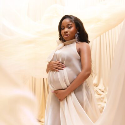 Fashion designer Veekee James in an ethereal cream silk maternity dress cradling her baby bump against a draped fabric backdrop.