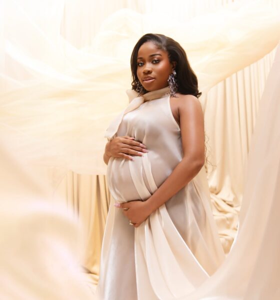 Fashion designer Veekee James in an ethereal cream silk maternity dress cradling her baby bump against a draped fabric backdrop.