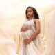 Fashion designer Veekee James in an ethereal cream silk maternity dress cradling her baby bump against a draped fabric backdrop.