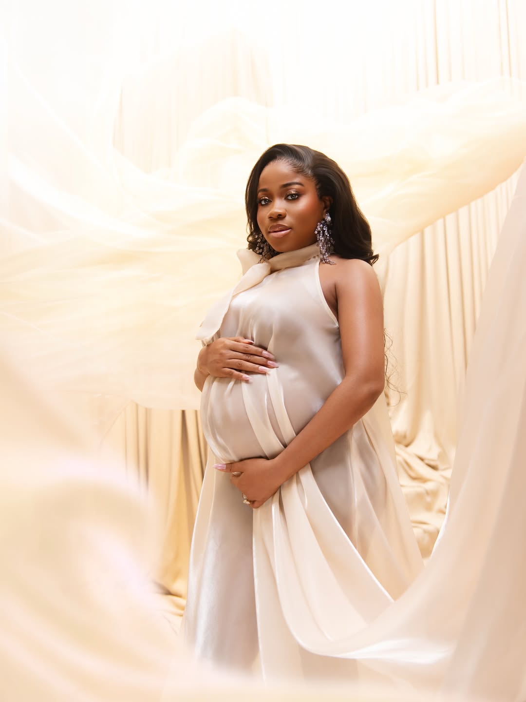 Fashion designer Veekee James in an ethereal cream silk maternity dress cradling her baby bump against a draped fabric backdrop.
