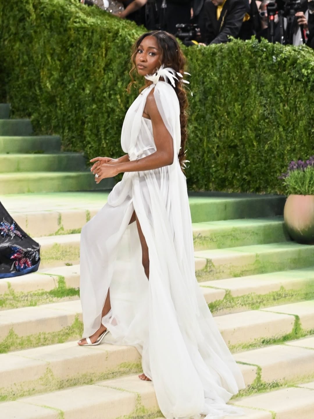 Ayo Edebiri ascending the steps at the 2026 Met Gala in an ethereal white chiffon Chanel gown by Matthieu Blazy with shoulder feather accents.