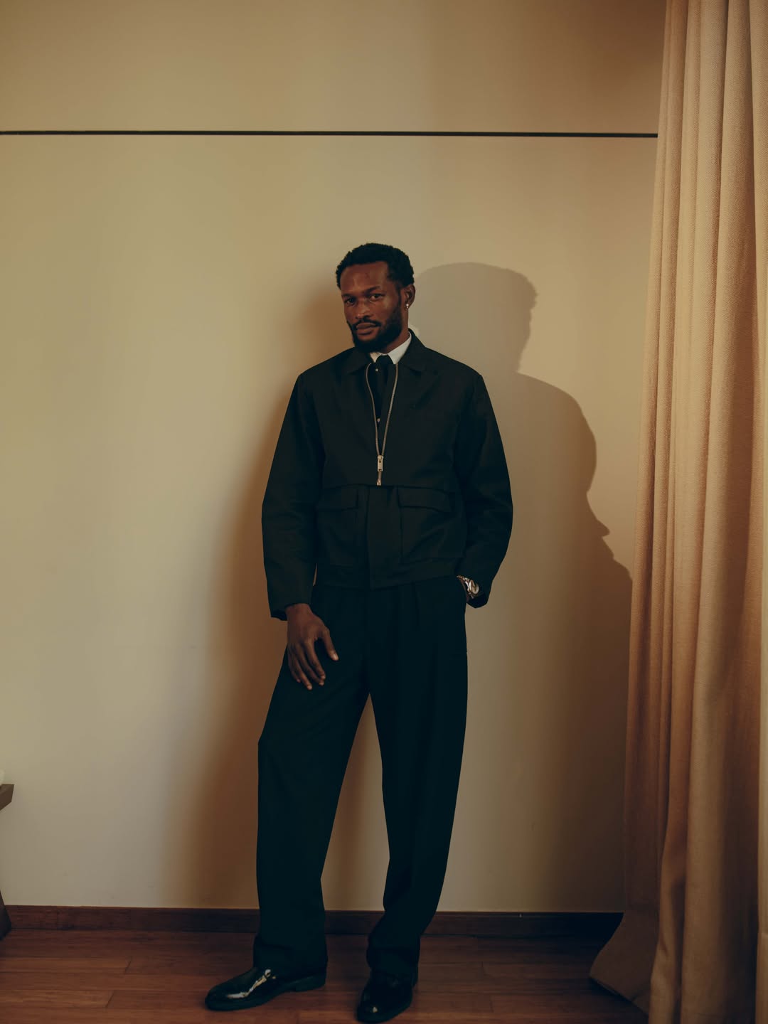 Nollywood actor Bucci Franklin standing against a neutral wall in a structured dark charcoal utility jacket, wide-leg trousers, and polished black shoes.