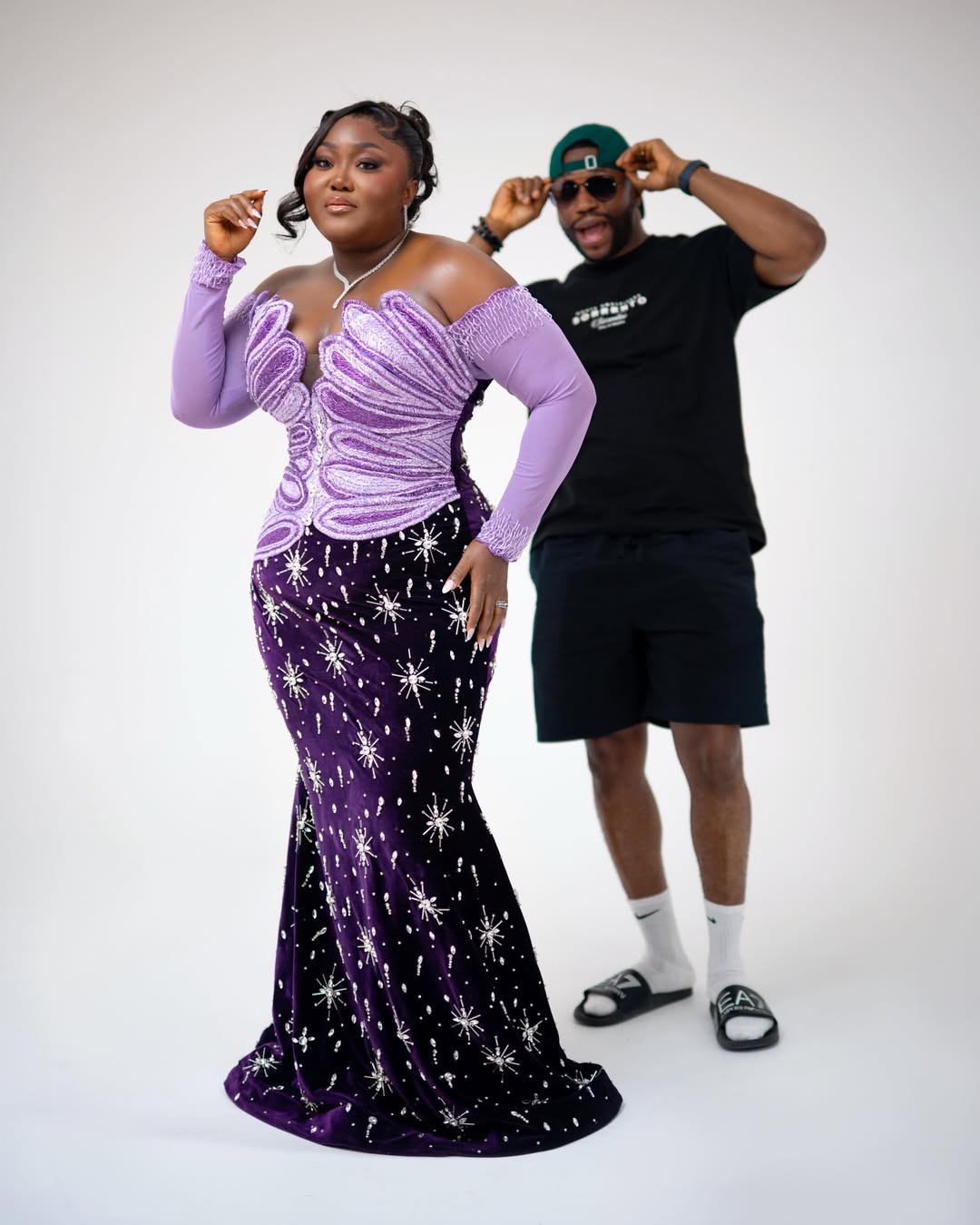Blessing Nze in a purple velvet mermaid gown with silver starburst embellishments, with husband Stan Nze visible in the background.