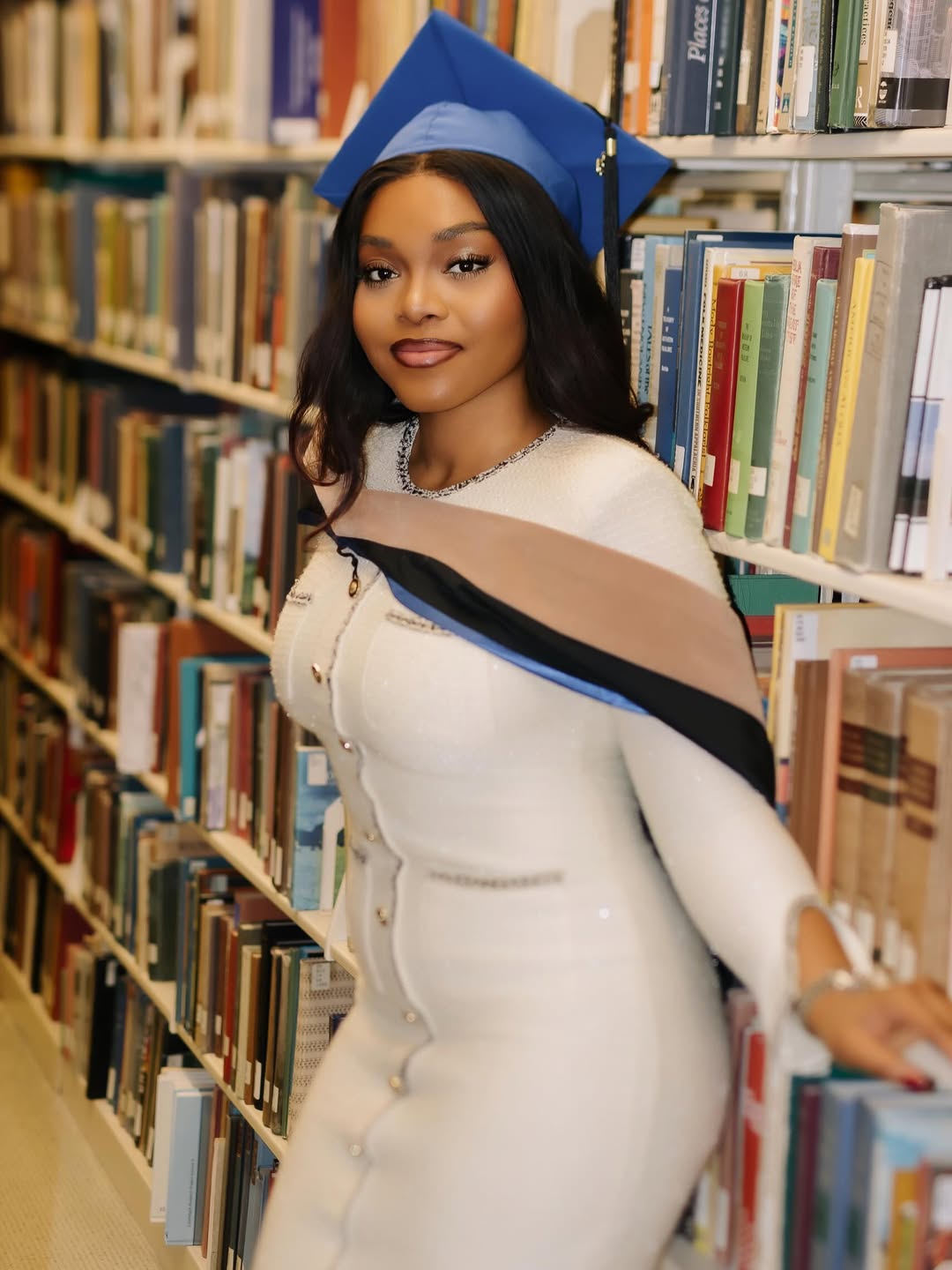 Lilo Aderogba leaning against a bookshelf in a library wearing her blue graduation cap and a white textured dress.