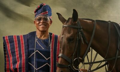 Farooq Oreagba standing majestically in a striped navy, crimson, and cream traditional Asọ-Ọkè Agbada next to a brown horse.