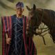 Farooq Oreagba standing majestically in a striped navy, crimson, and cream traditional Asọ-Ọkè Agbada next to a brown horse.