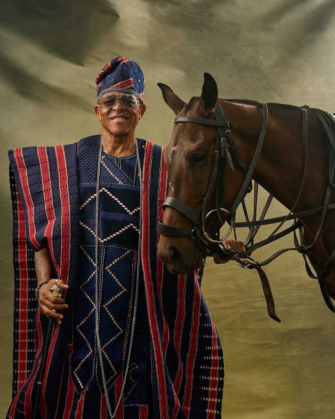 Farooq Oreagba standing majestically in a striped navy, crimson, and cream traditional Asọ-Ọkè Agbada next to a brown horse.