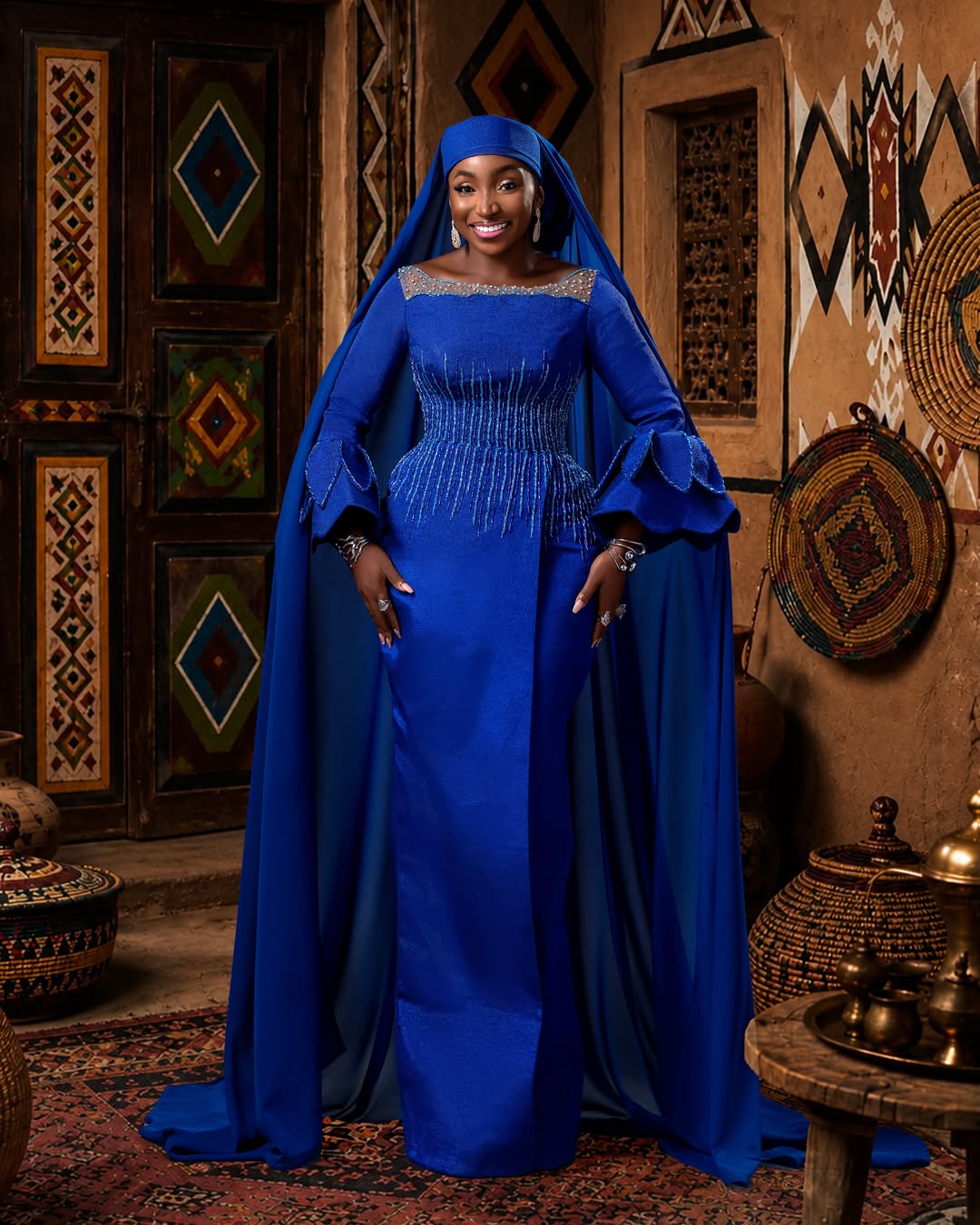 Full-length portrait of Omawunmi Dada wearing a structured royal blue column gown with tiered sleeve ruffles and a matching headpiece for the Okanjuwa movie premiere.