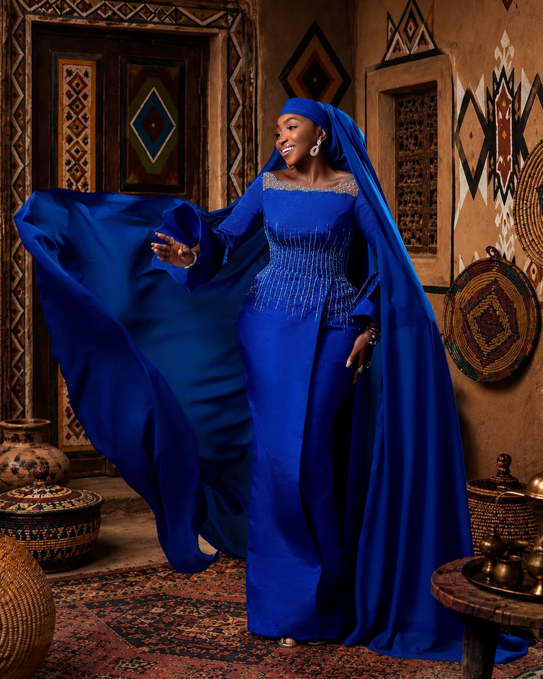 Actress Omawunmi Dada posing with a flowing royal blue chiffon veil cape in motion against a traditional Hausa architectural backdrop for the Okanjuwa film premiere.