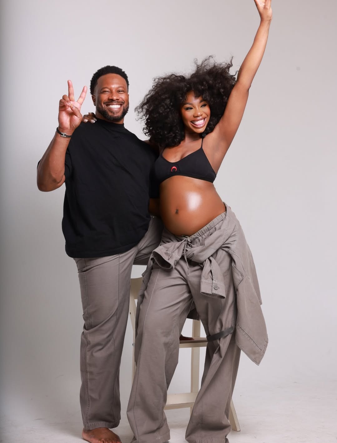 Sharon Ooja and husband Ugo Nwoke celebrate during her maternity shoot, Sharon raising one arm in joy while Ugo flashes a peace sign, both wearing matching grey and black outfits