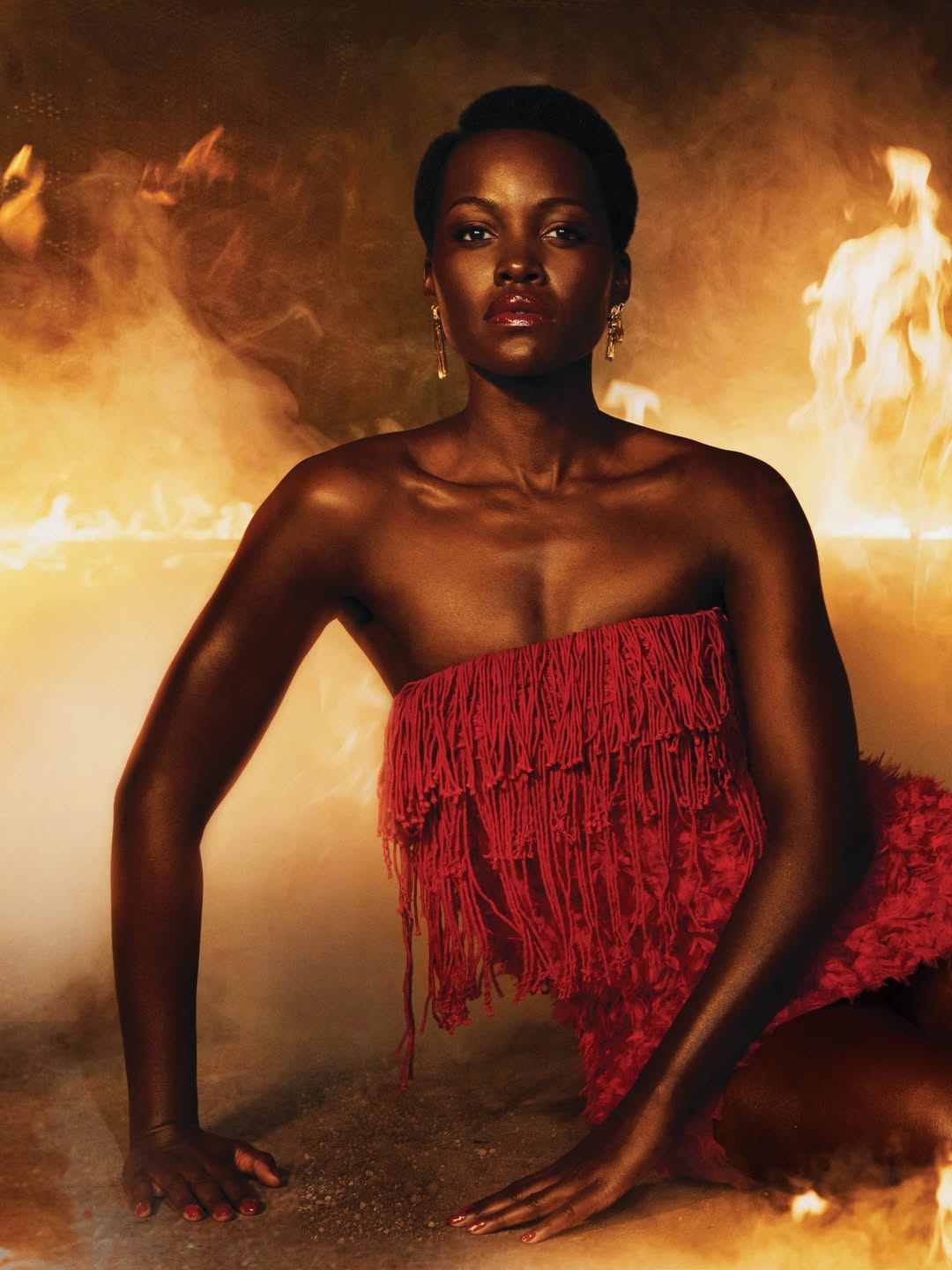 Close-up of Lupita Nyong'o wearing a red strapless fringe mini dress from Mugler during the ELLE Magazine 2026 "The Odyssey" promotional shoot with dramatic smoke and fire.