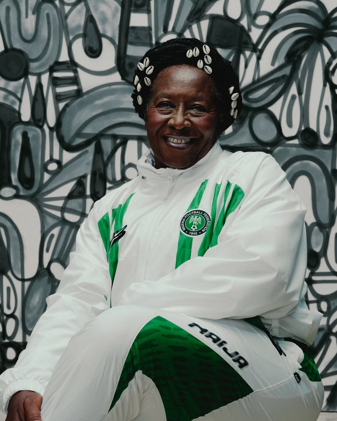 Close-up portrait of actress Patience Ozokwo smiling whilst wearing a white Nike zip-up tracksuit jacket with vertical green stripes and the Nigeria Football Federation crest, accessorised with cowrie shells in her hair.