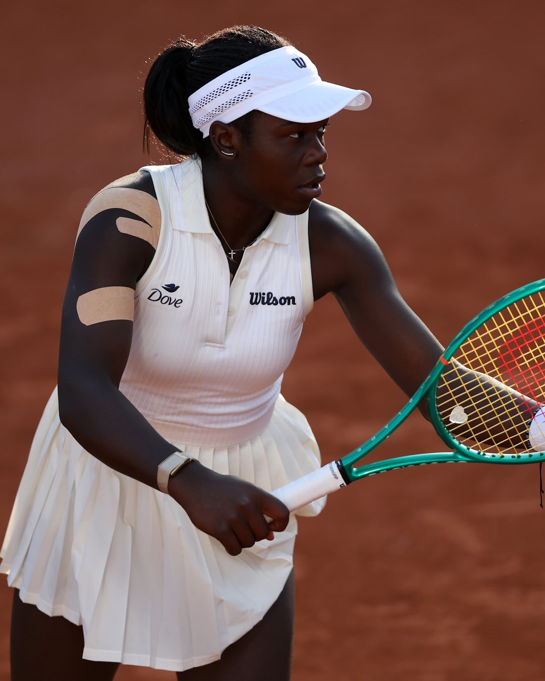 Close-up of a focused Victoria Mboko holding her tennis racket in a ready position, wearing a white Wilson visor and tennis dress at the 2026 French Open.