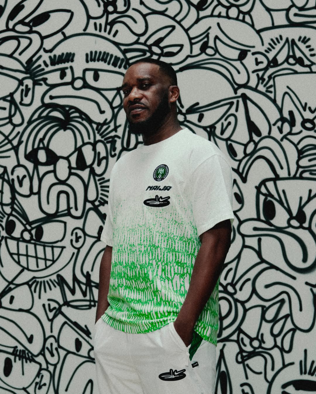 Football legend Jay-Jay Okocha standing with hands in pockets, wearing a white relaxed-fit Nike t-shirt with a spray-gradient lime green bottom half, the NFF crest, and Slawn's custom "NAIJA" script logo.