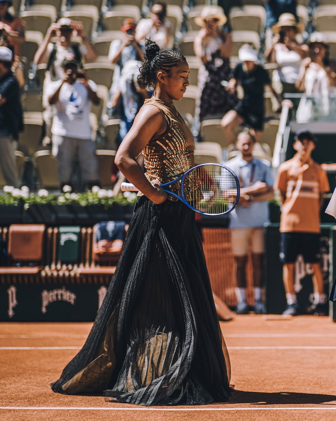 Naomi Osaka stands on Court Suzanne-Lenglen holding her racket in a gold sequined Nike dress and black Kevin Germanier tulle skirt during her first-round French Open 2026 match against Laura Siegemund on 26 May
