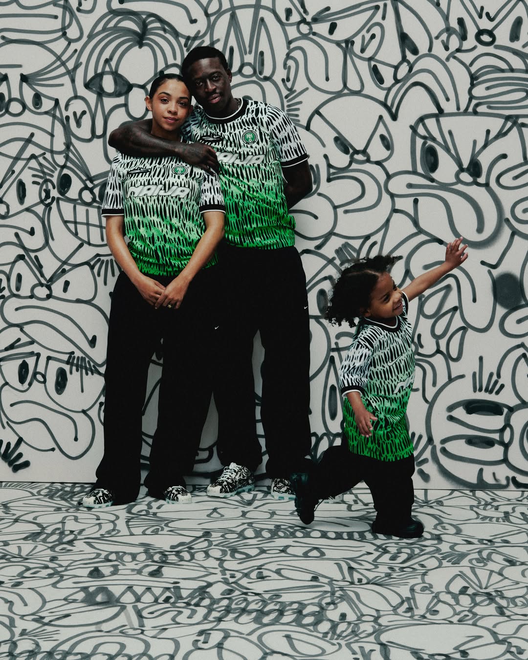 London-based artist Slawn posing with his partner and child against a black-and-white cartoon doodle wall, with all three wearing matching green and white patterned Nike jerseys from his 2026 collaboration.