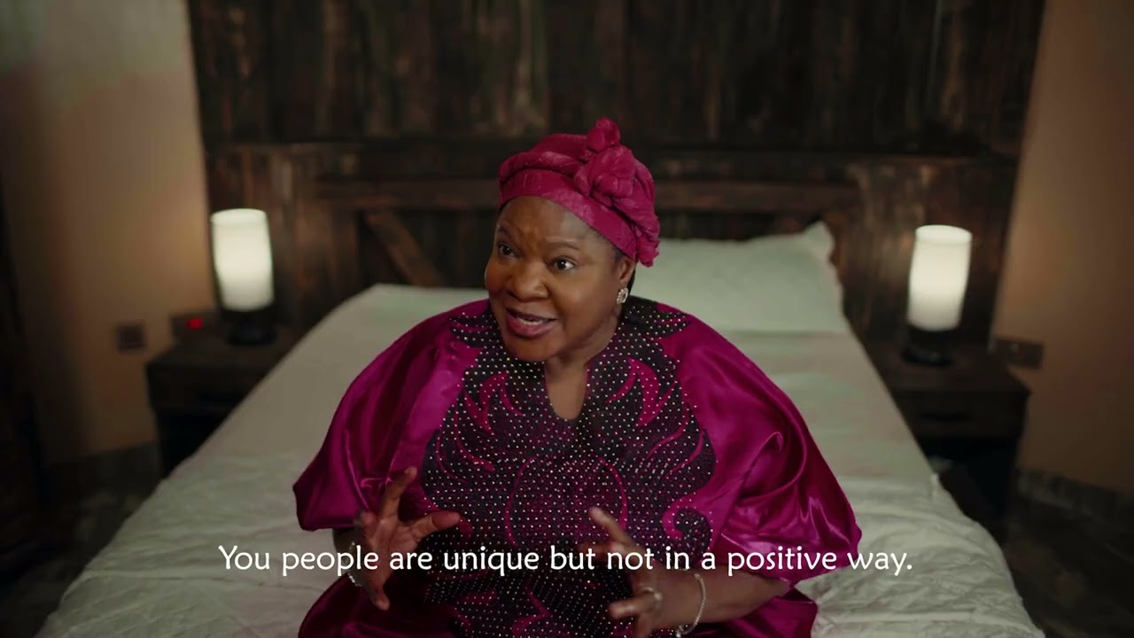A movie character still from Ajosepo 2: The Gathering showing actress Toyin Abraham in a vibrant magenta traditional outfit speaking expressively on a bed,