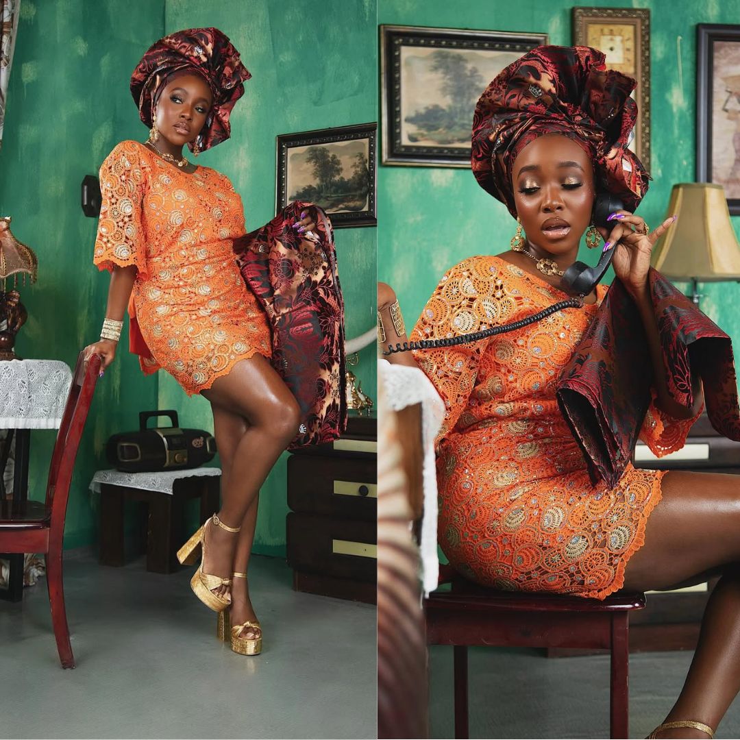 Actress Ini Dima-Okojie in a vibrant orange lace Oleku paired with a dark red patterned gele, posing next to a vintage telephone.